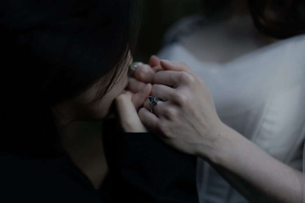 Close-up of two people holding hands tenderly in a dark, intimate setting, one wearing a silver ring.