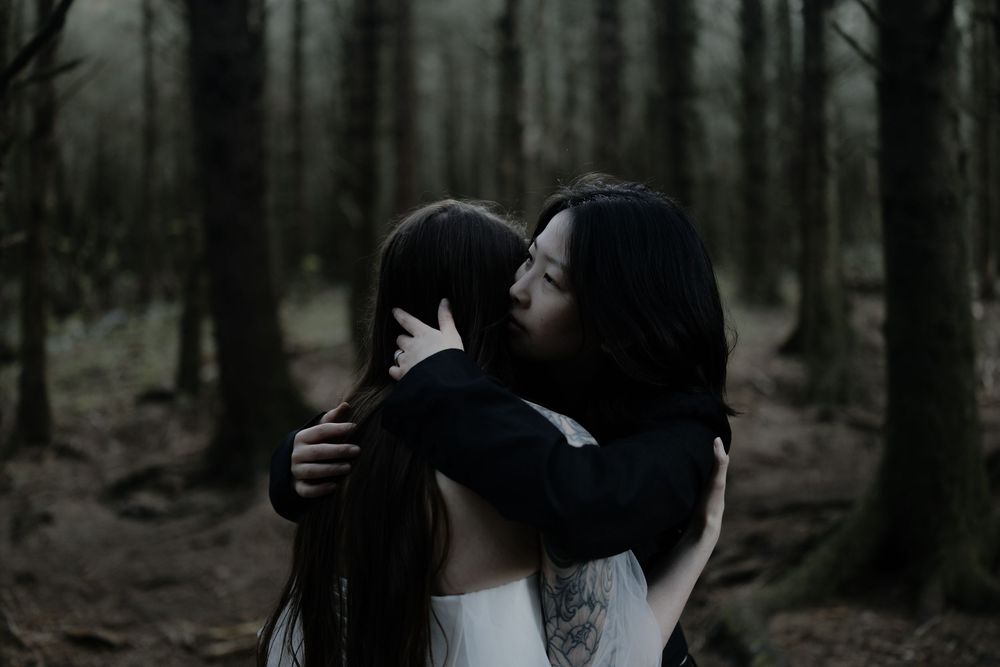 Two women embracing in a dark, moody forest setting, sharing an emotional hug among tall trees.