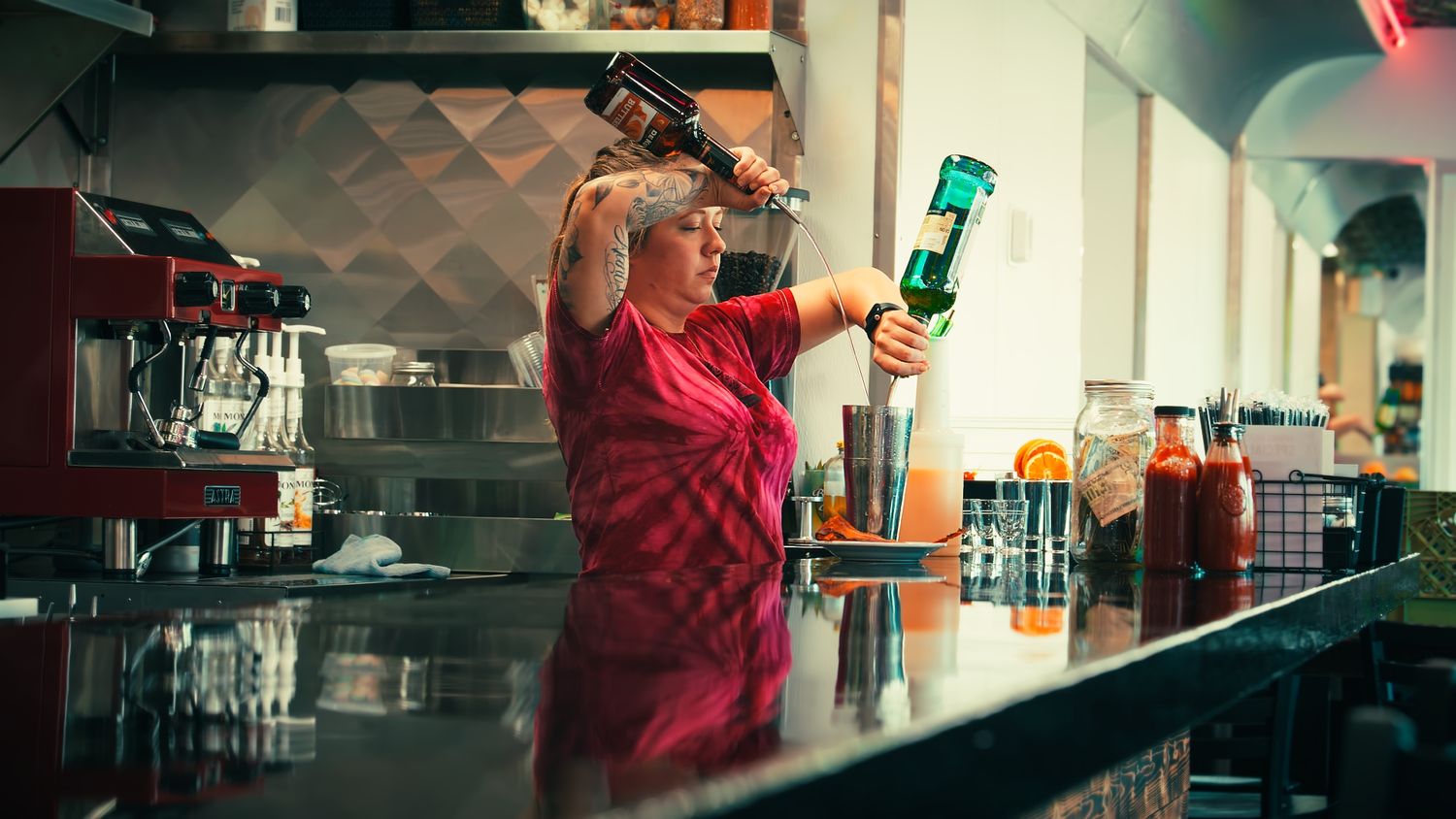 Female bartender pouring drinks behind a bar counter in a colorful restaurant setting.