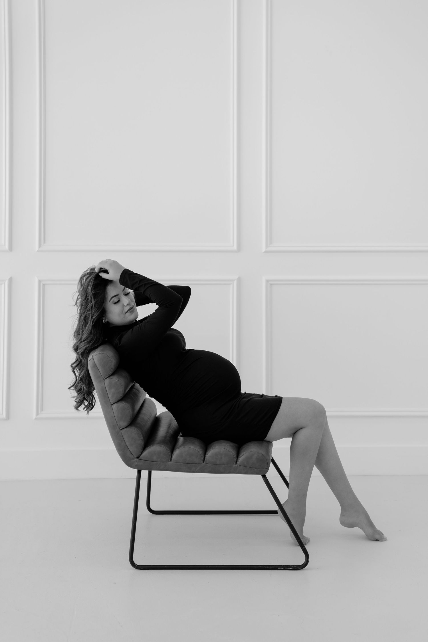 Pregnant woman in black turtleneck dress posing elegantly beside a brown leather chair in a white room.