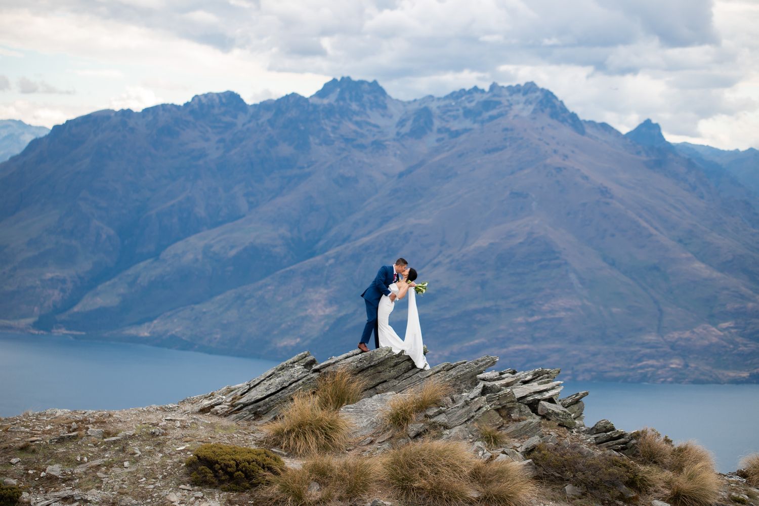 Couple stands atop rocky outcrop overlooking scenic mountain lake and rugged peaks in New Zealand.