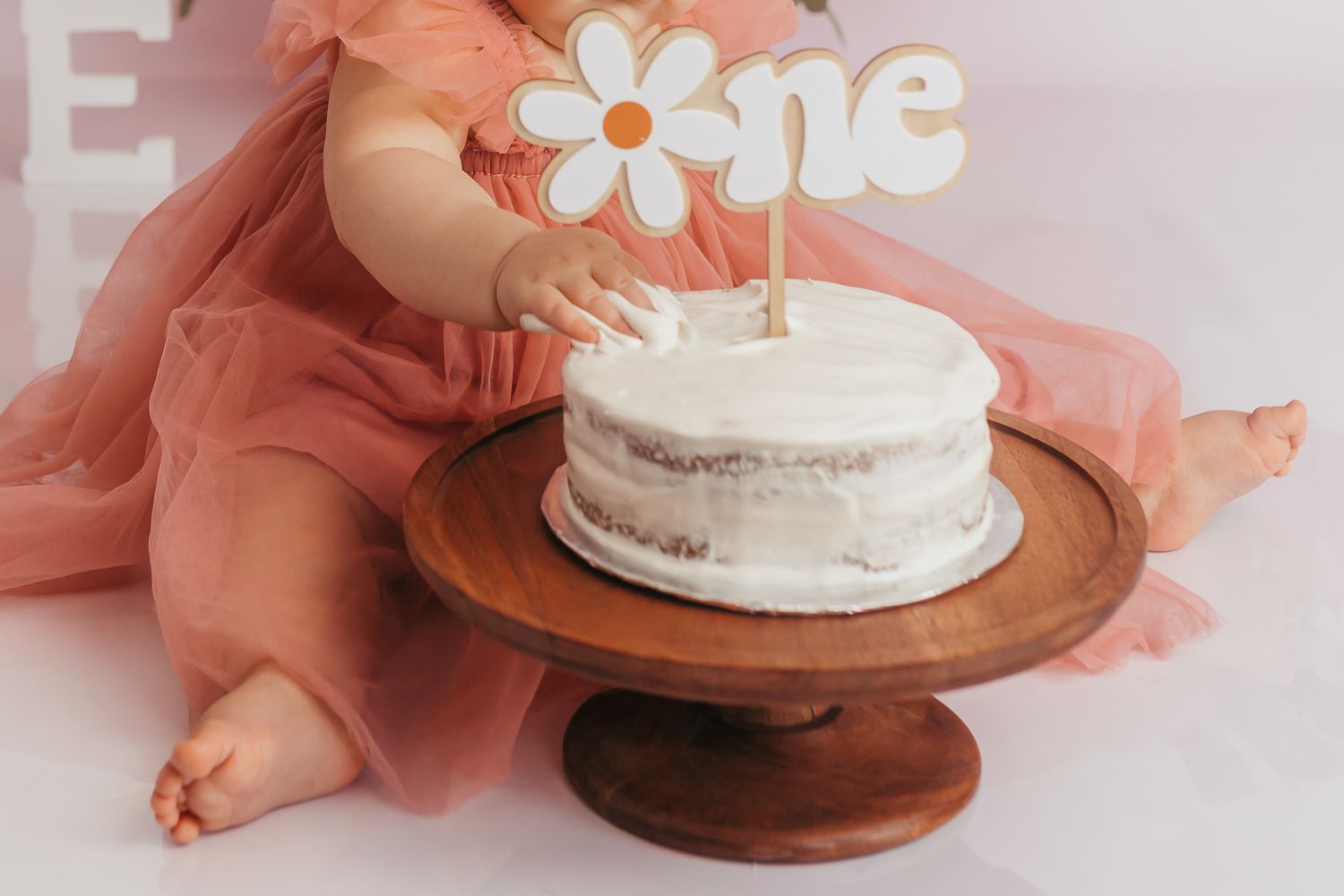 Baby in pink tulle dress touching a white smash cake on wooden stand with a daisy \"one\" topper for first birthday.