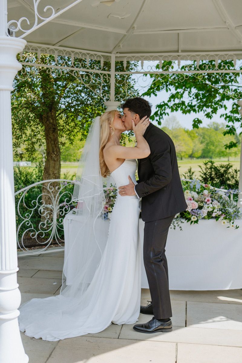 Bride and groom share first kiss under white gazebo adorned with flowers at outdoor wedding ceremony.
