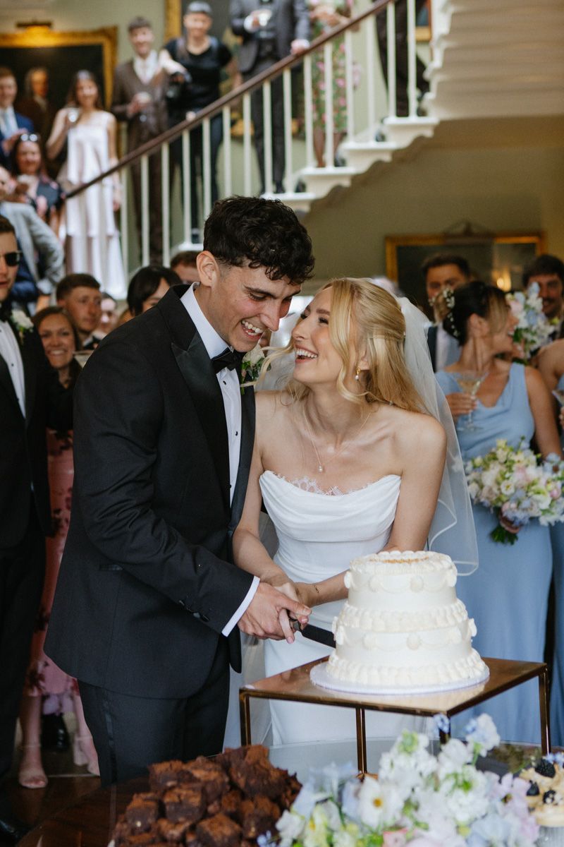Bride and groom smiling while cutting their white tiered wedding cake surrounded by guests at an elegant venue.