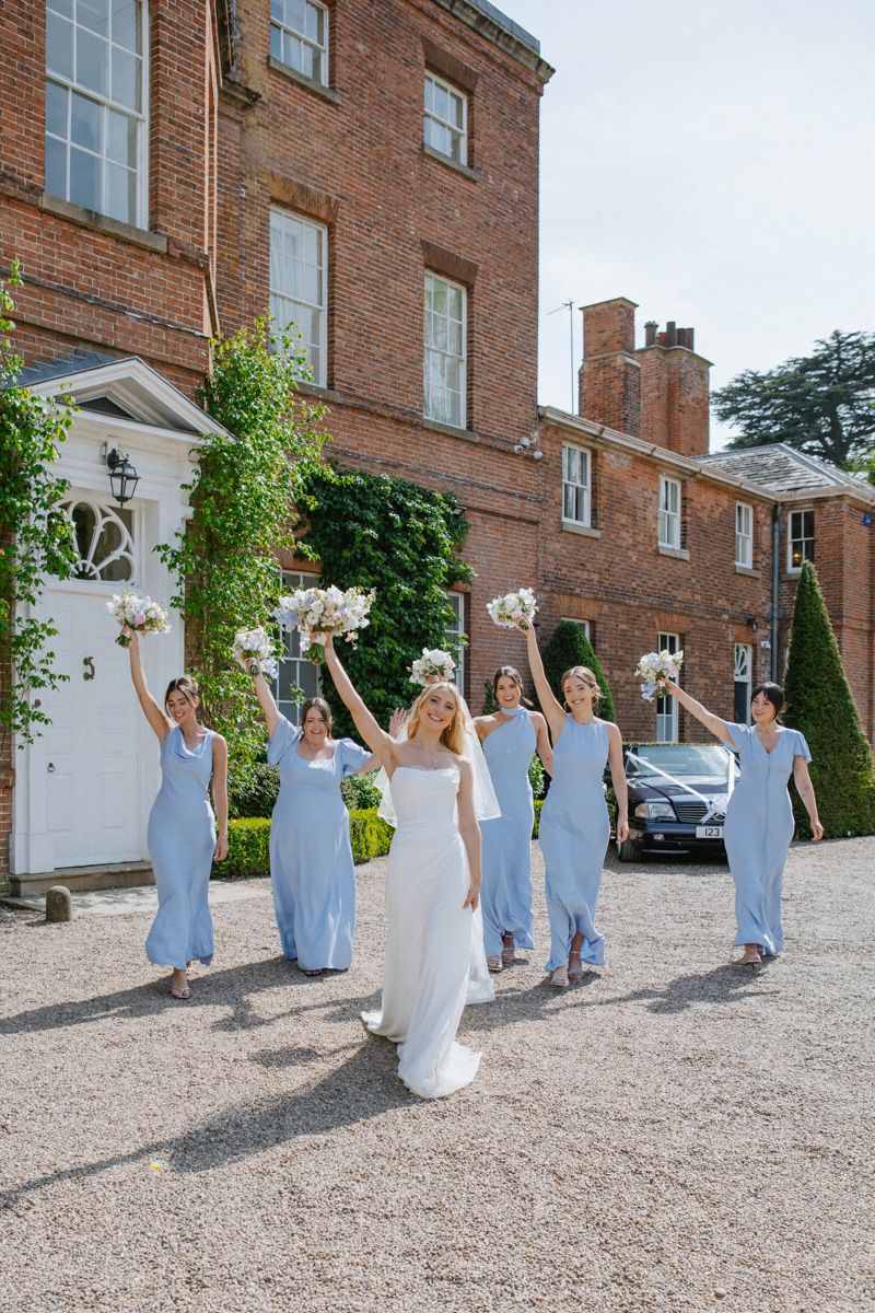 Bride in white gown leads bridesmaids in blue dresses raising bouquets outside elegant brick manor.