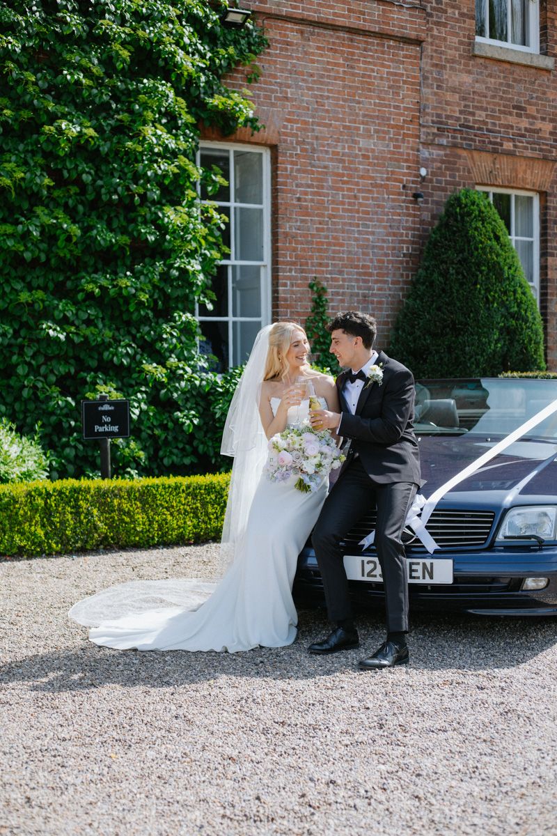A bride and groom pose together leaning against a decorated wedding car outside a brick manor house.