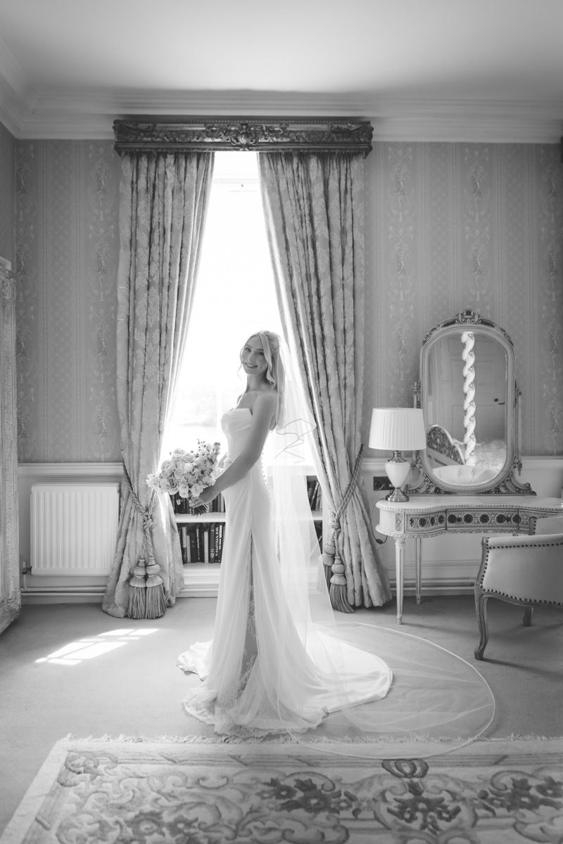 Elegant bride in white gown with train stands by window in ornate room with vintage mirror and drapes, black and white photo.