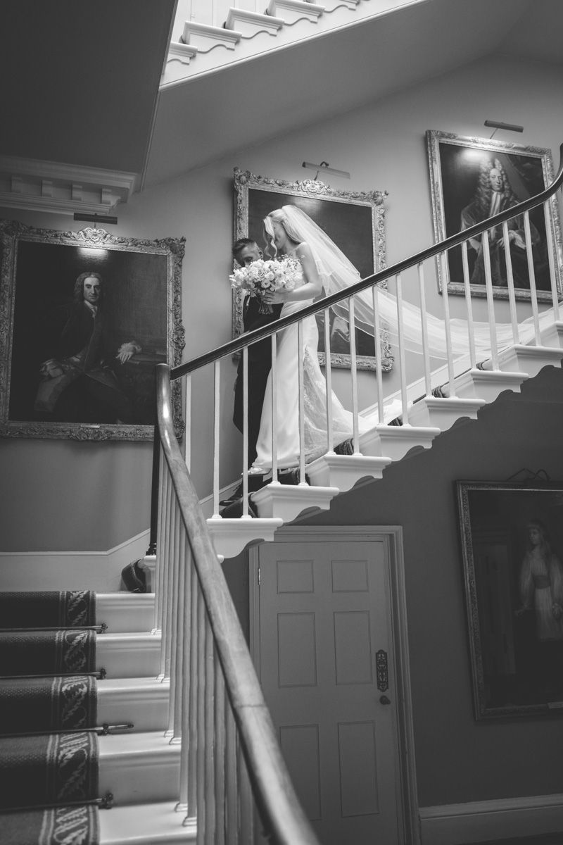 Black and white photo of a bride in a flowing gown and veil descending an elegant staircase adorned with portrait paintings.