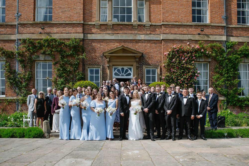 Large wedding party posing outside a grand brick manor house with bridesmaids in blue dresses.