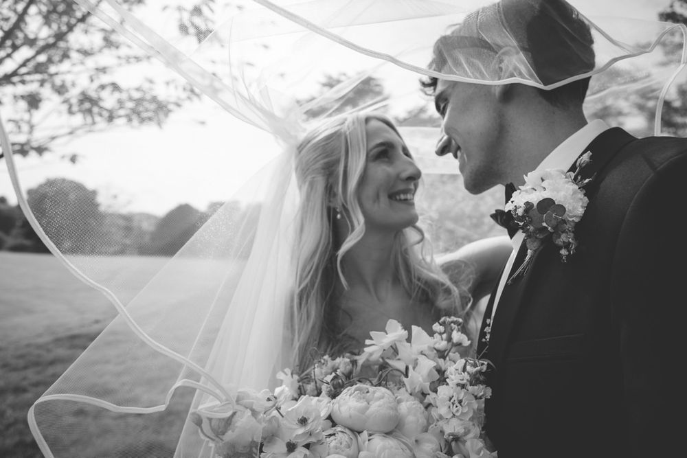 Black and white wedding photo of smiling bride with veil and bouquet gazing at groom in suit outdoors.