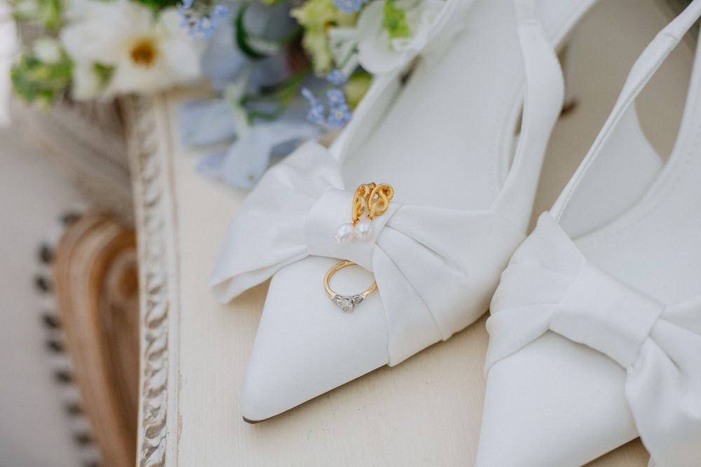 White bridal heels with bow detail and diamond engagement ring displayed on elegant surface with floral bouquet.
