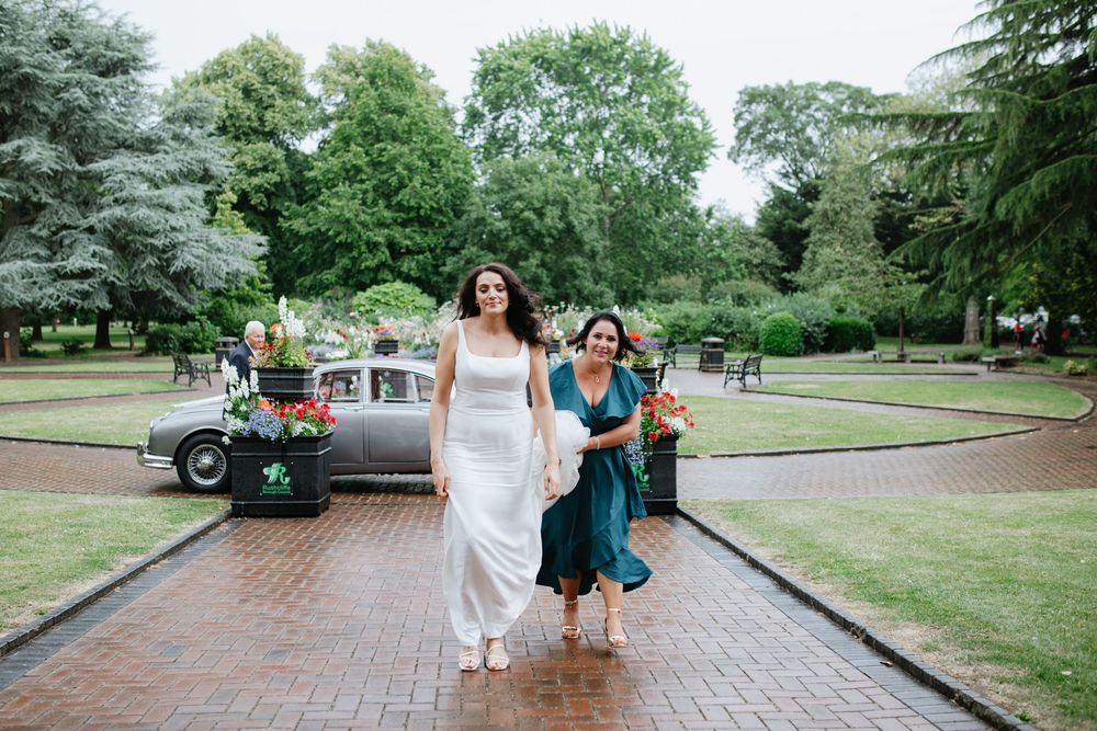 Bride in white gown walks with bridesmaid in teal dress along brick path in lush green park setting.