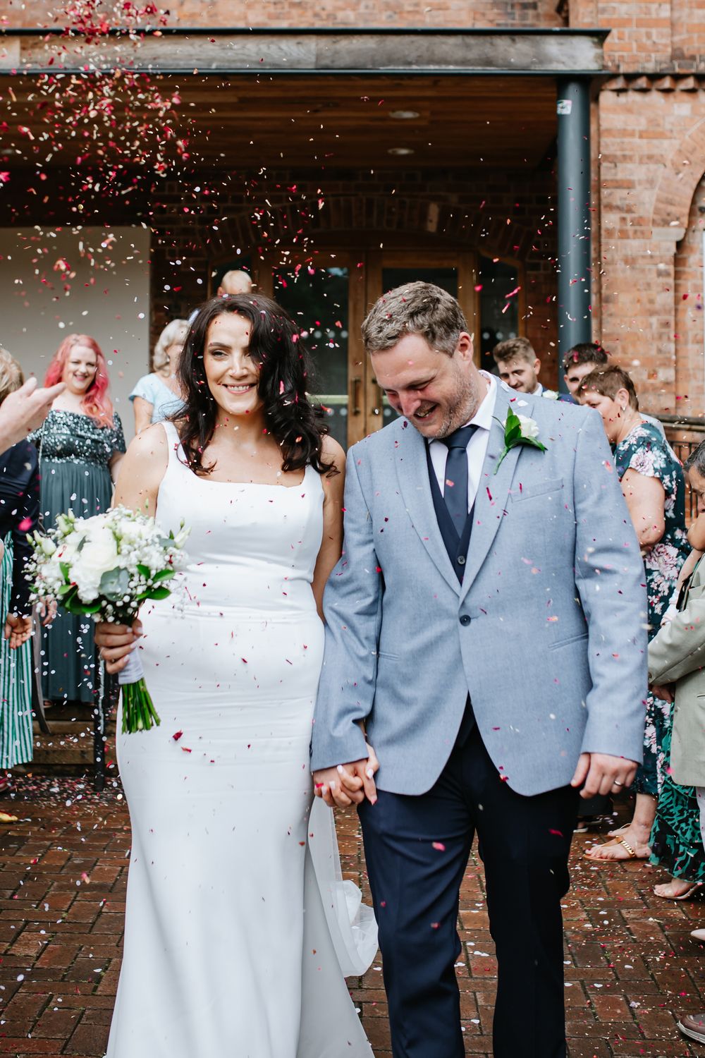 Bride and groom walk out of wedding venue holding hands as colorful confetti falls around them.