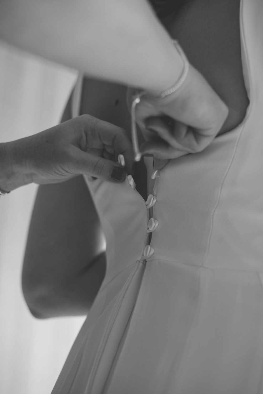 Black and white photo of hands fastening small buttons on the back of a white wedding dress.