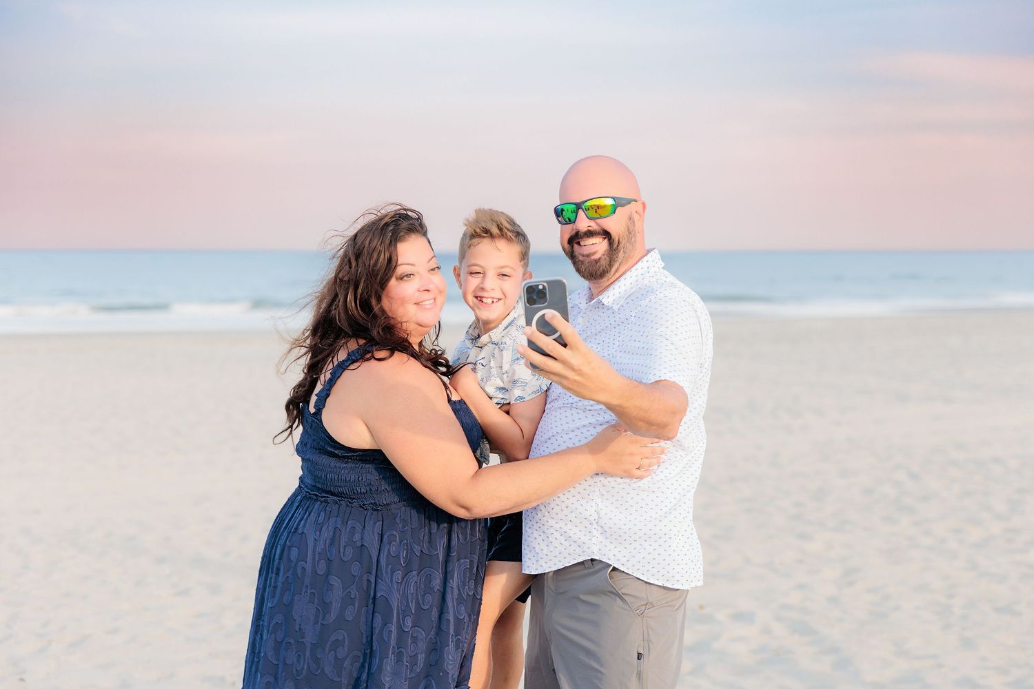 Family of three smiling on a sandy beach at sunset, parents holding young child, wearing casual summer attire.