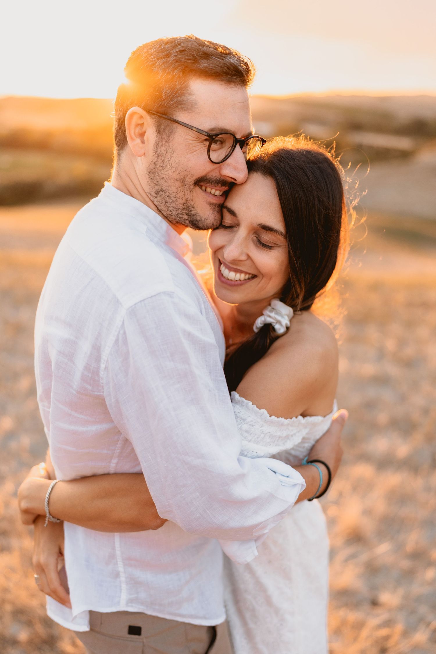 Couple embracing at golden sunset in a field, man in white linen shirt with glasses, woman in white off-shoulder dress smiling.