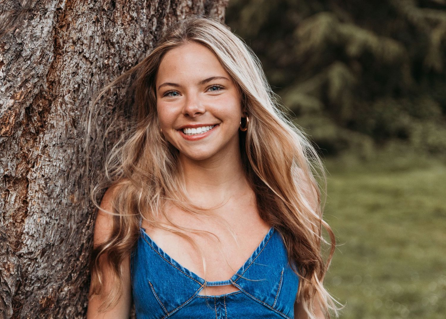 Smiling young woman with long blonde hair leaning against a tree in a denim top outdoors.