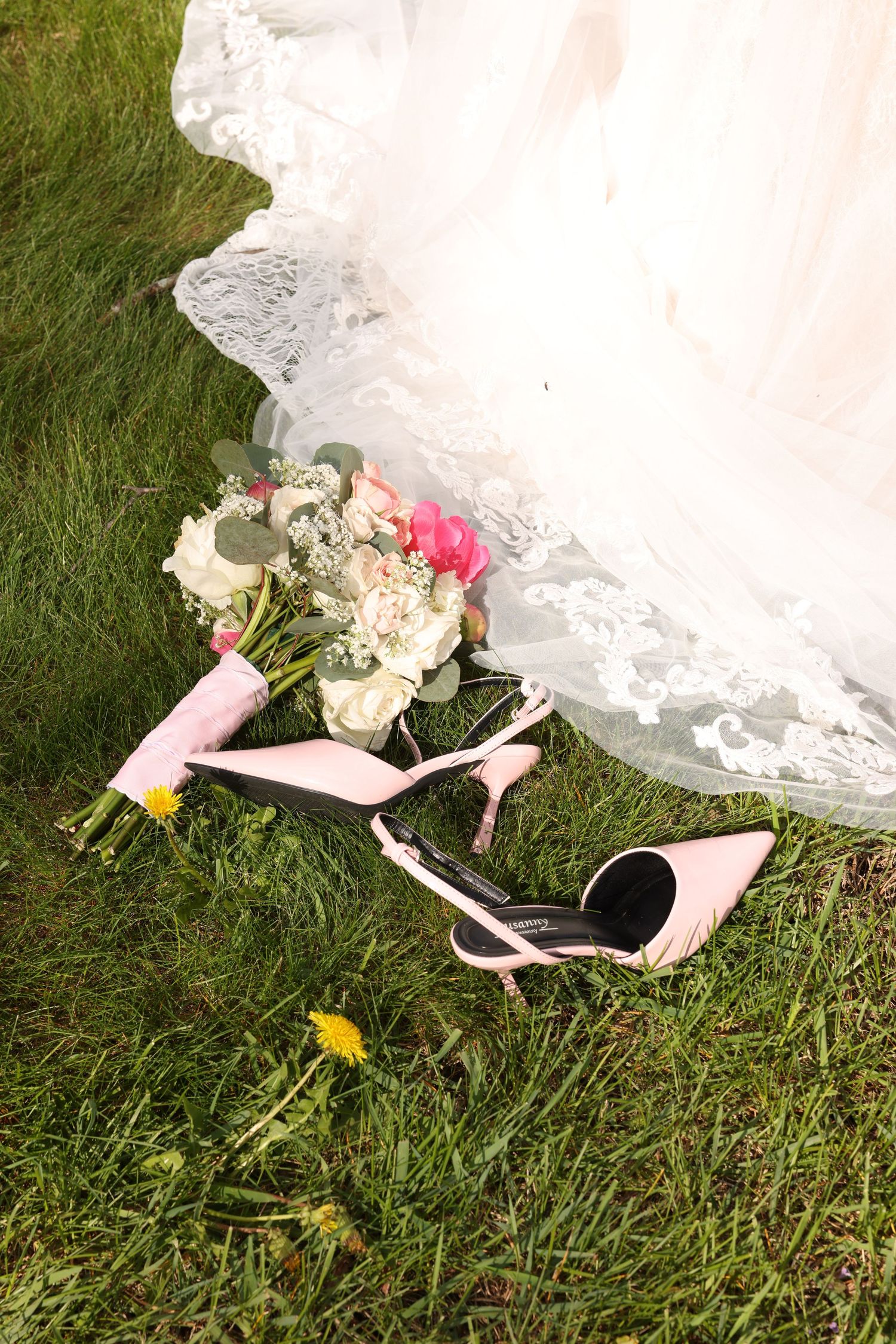 Pink bridal heels and floral bouquet resting on green grass beside a white lace wedding dress hem.