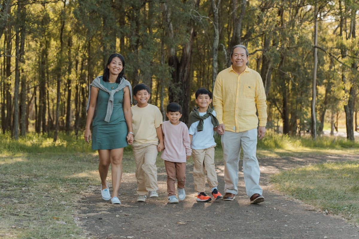 Asian family of five walking together on a sunny forest path, smiling and holding hands outdoors in sydney during a rouse hill photoshoot