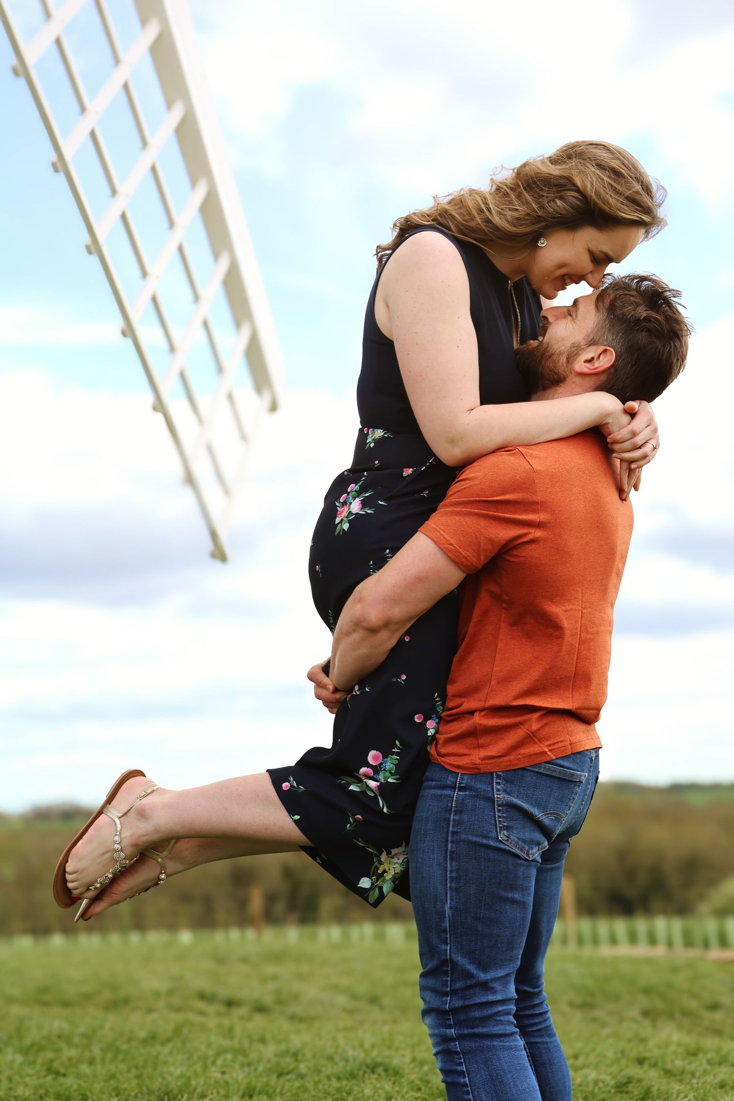 A man in an orange shirt lifts a woman in a floral dress near a windmill on a green field.