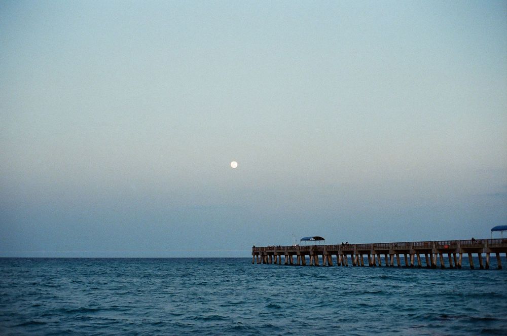A wooden pier extends over calm blue ocean waters under a hazy dusk sky with a full moon rising.