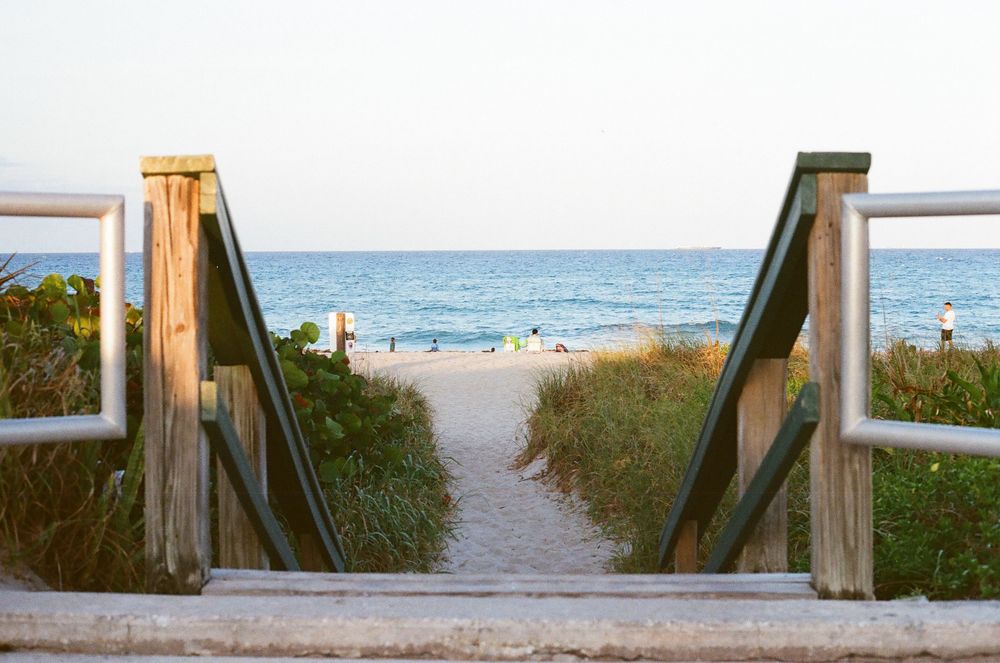 Wooden boardwalk path leads through sea grass to a beautiful sandy beach with calm blue ocean waters.