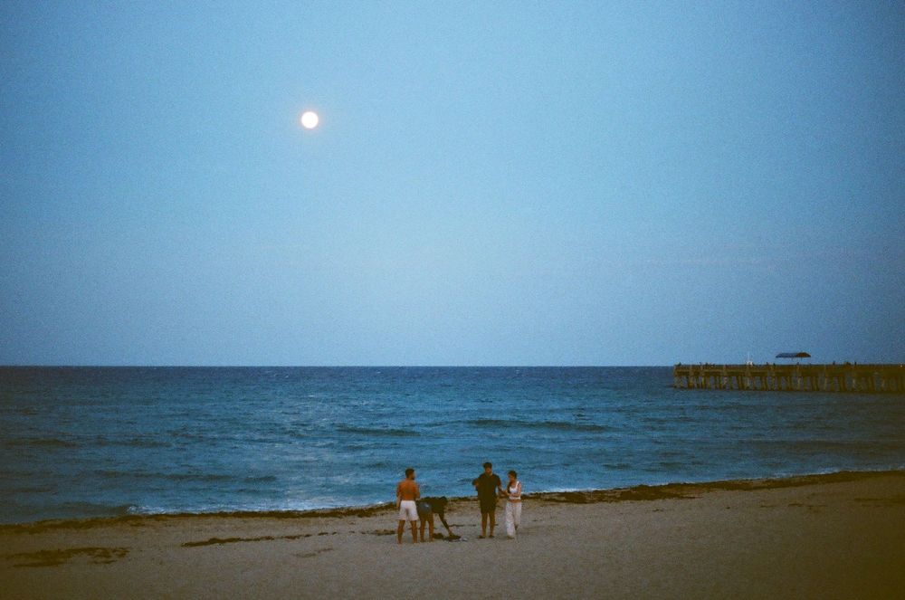Family with dog standing on sandy beach at dusk with full moon rising over calm ocean near pier.