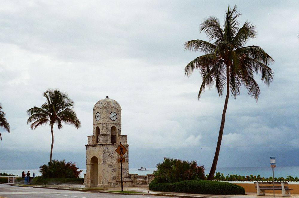 Historic clock tower landmark flanked by tall palm trees under dramatic cloudy skies in Palm Beach Florida.