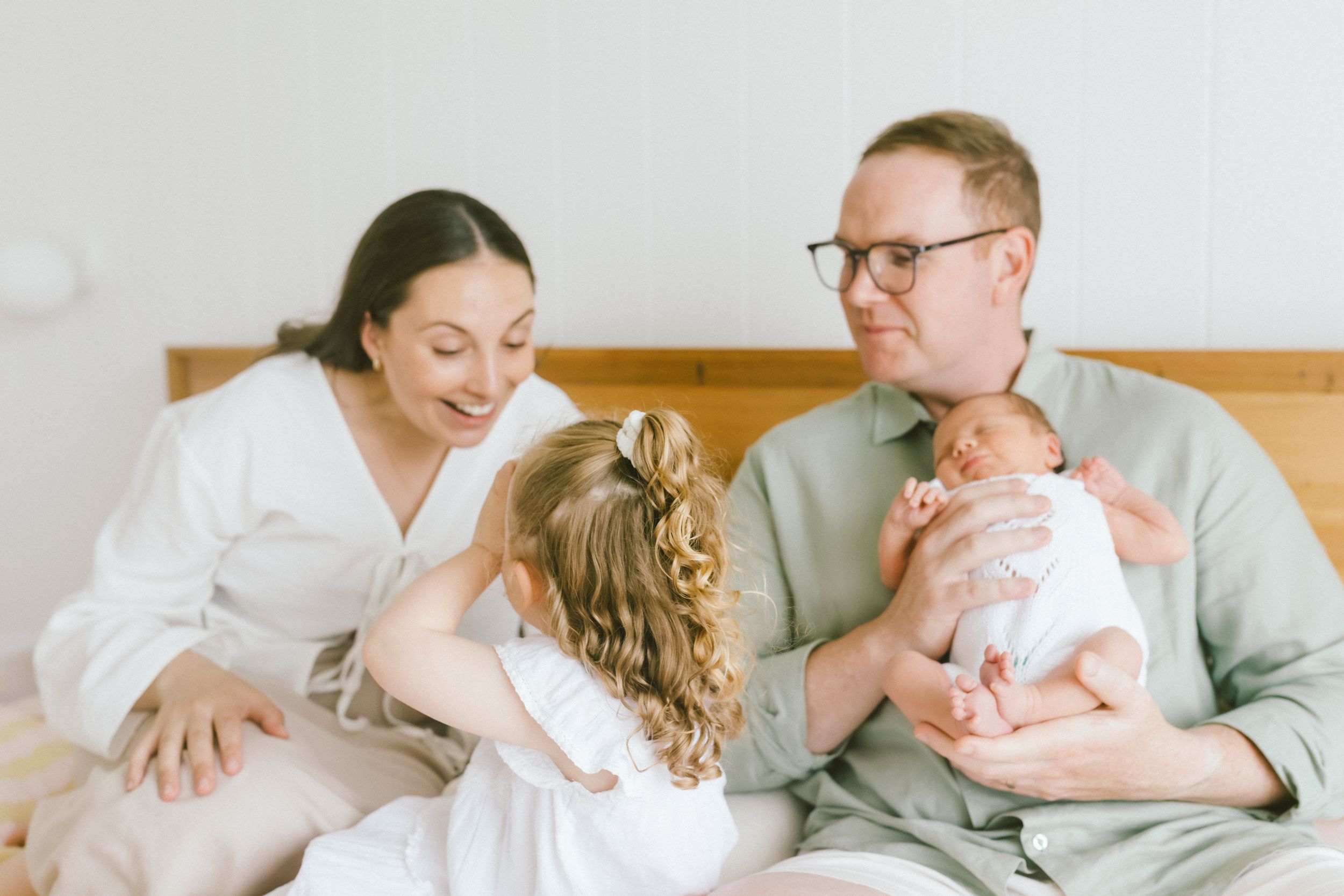 Family of four sitting on bed, parents smiling at newborn baby while young child reaches out to touch sibling.