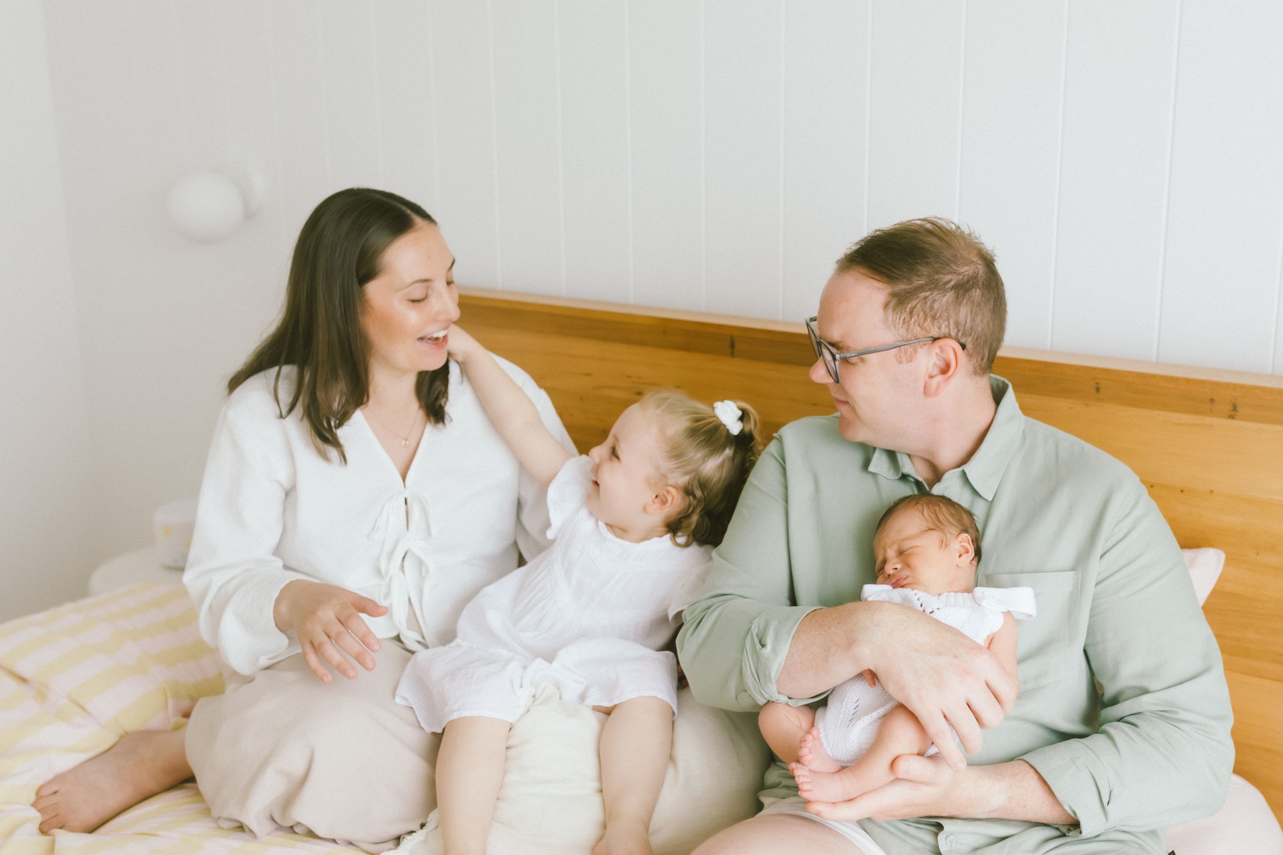 Happy family of four sitting on bed, parents smiling at toddler girl while dad holds newborn baby.