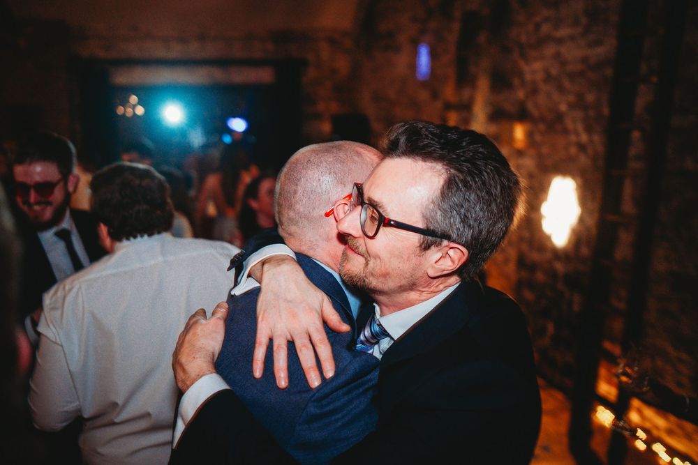 Two men share a warm embrace at a wedding reception in a rustic venue with dim atmospheric lighting.