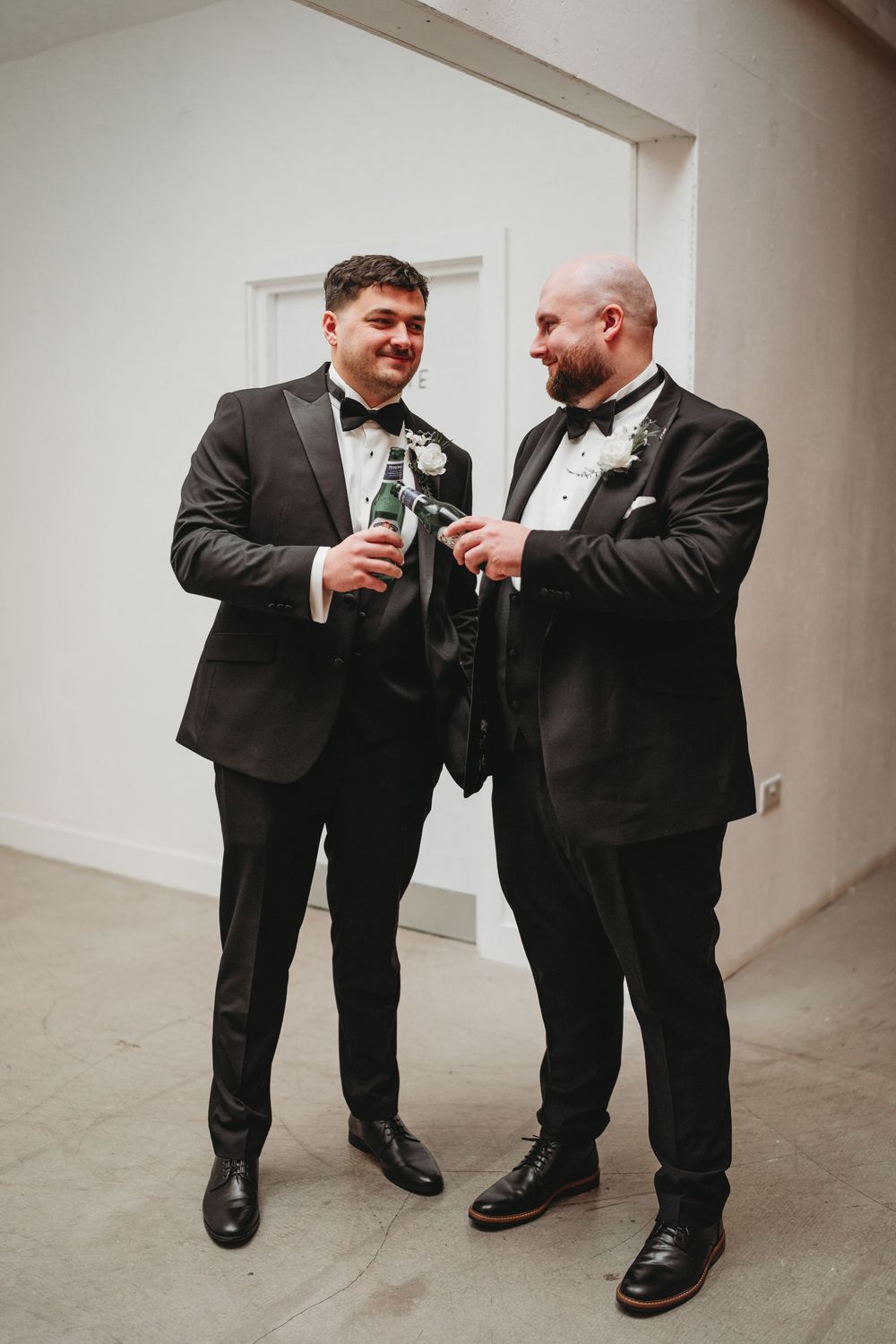 Two grooms in black tuxedos clinking beer bottles and smiling at each other during their wedding celebration.