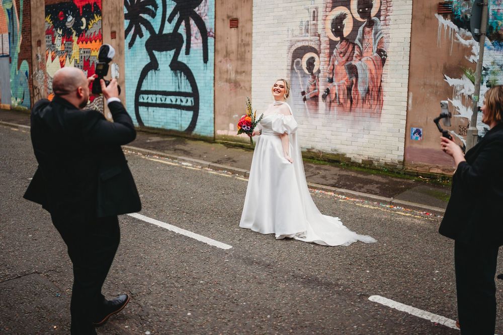 Bride in white gown poses before colorful street art mural while guests photograph her.