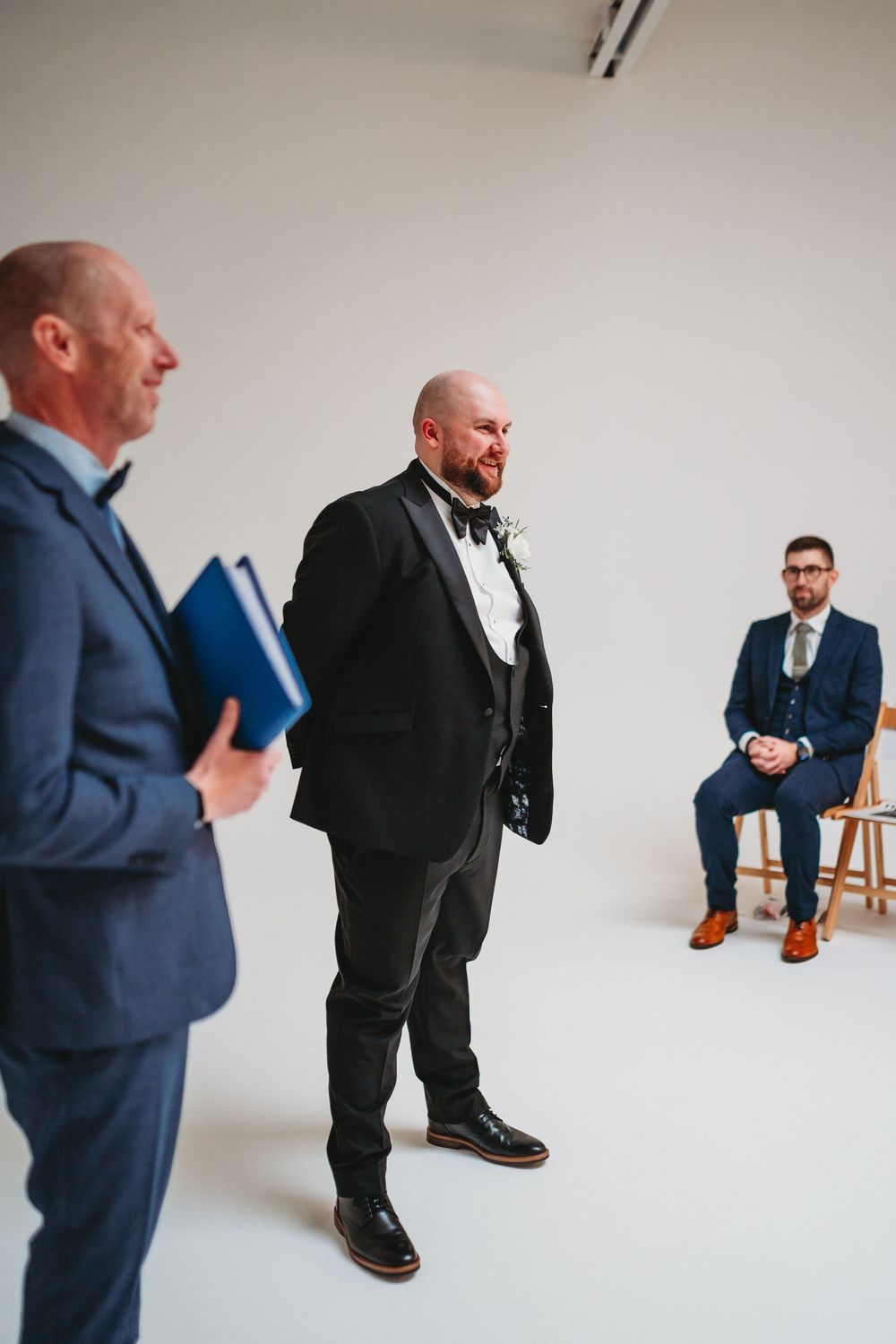 Bald groom in black tuxedo with bow tie waiting at altar during wedding ceremony.