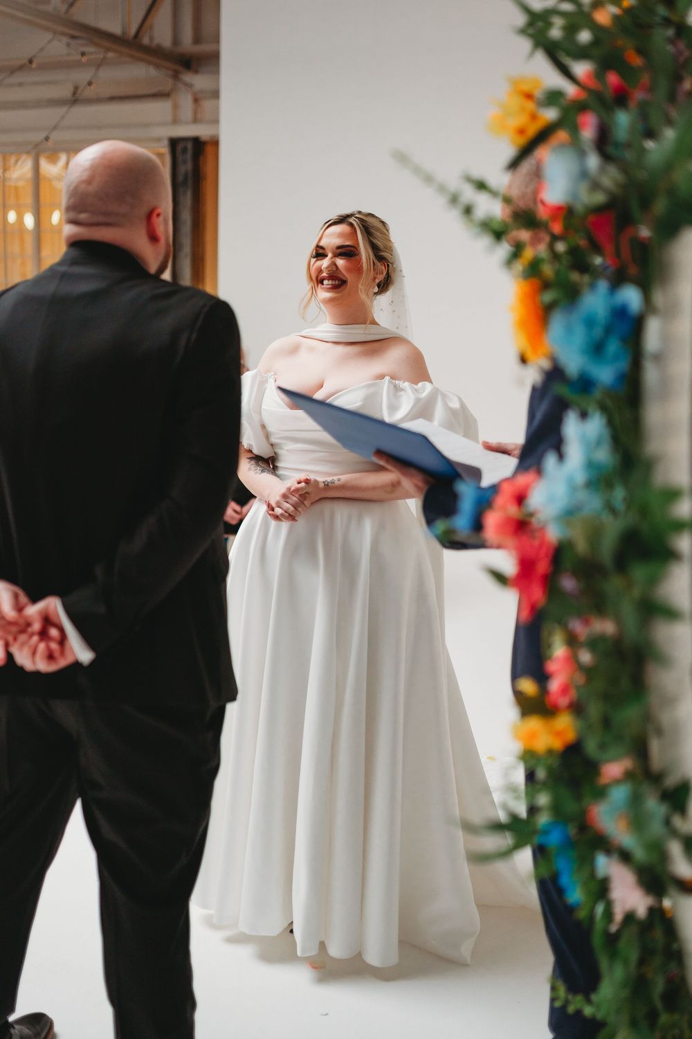 Smiling bride in off-shoulder white gown reads vows at colorful floral arch wedding ceremony.