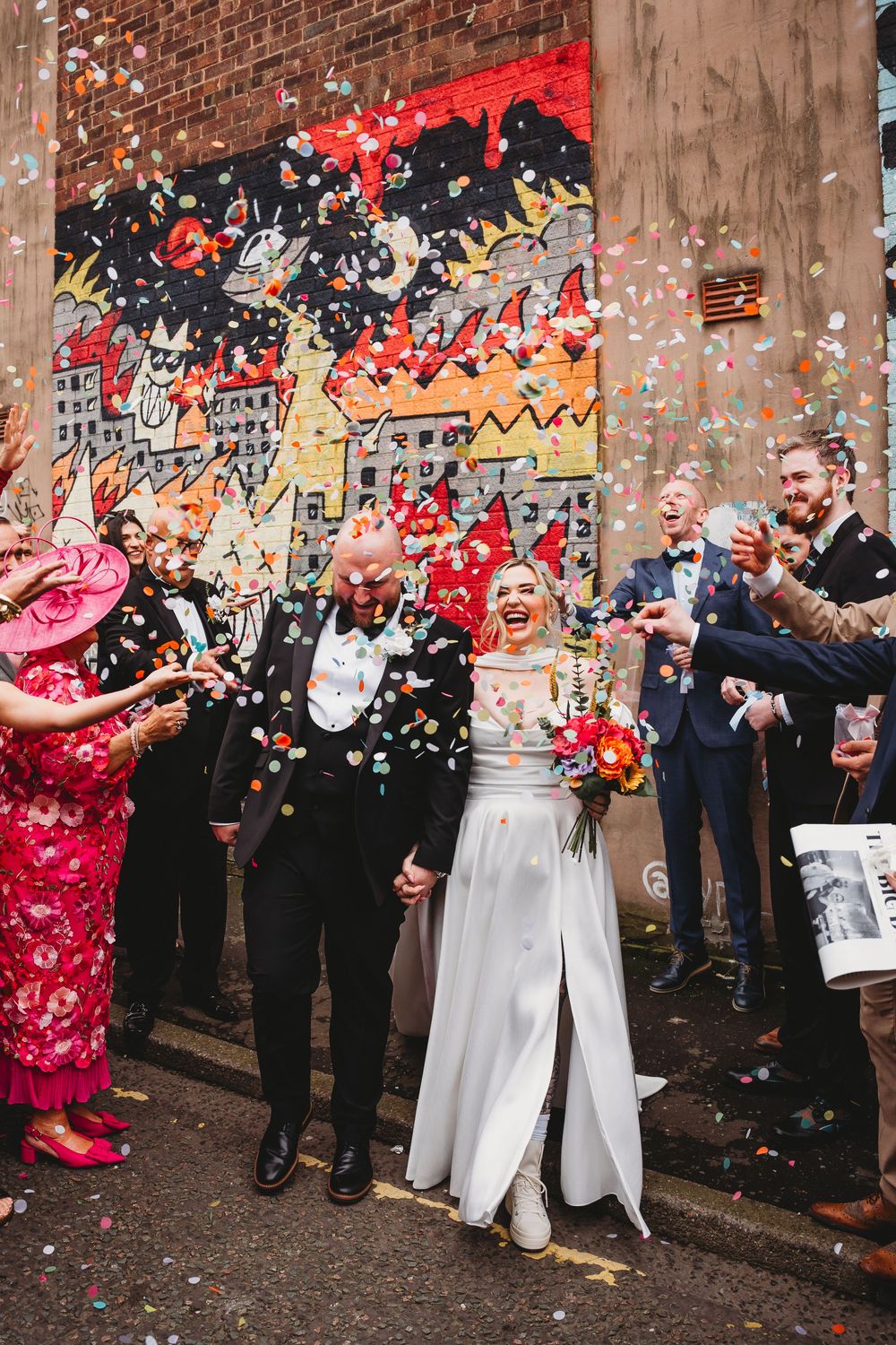 Newlyweds celebrate with colorful confetti shower outside brick wall with vibrant street art mural.
