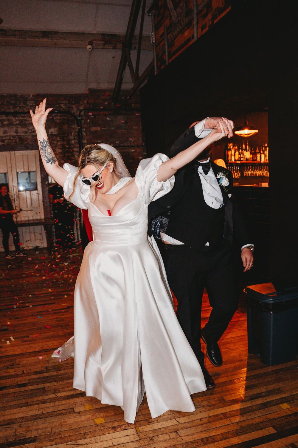 Bride in sunglasses and groom dance energetically at indoor wedding reception.