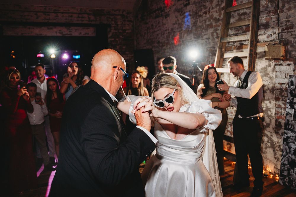 Bride in white gown wearing sunglasses dances with tuxedo-clad guest at lively wedding reception.