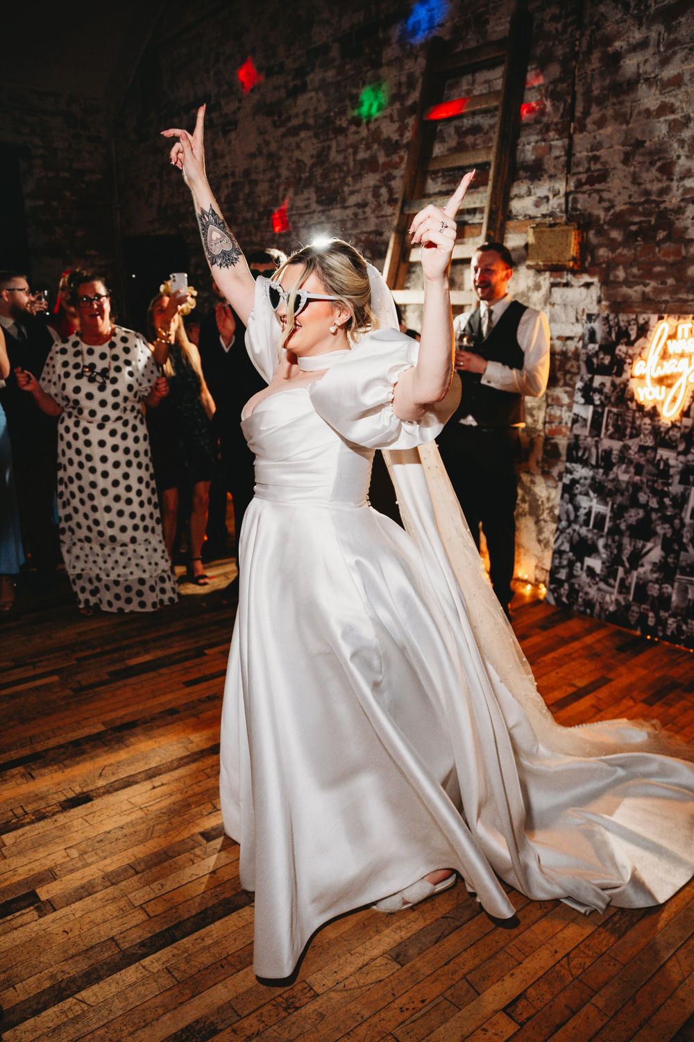 Bride in white satin gown and sunglasses dancing joyfully on wooden dance floor at wedding reception.