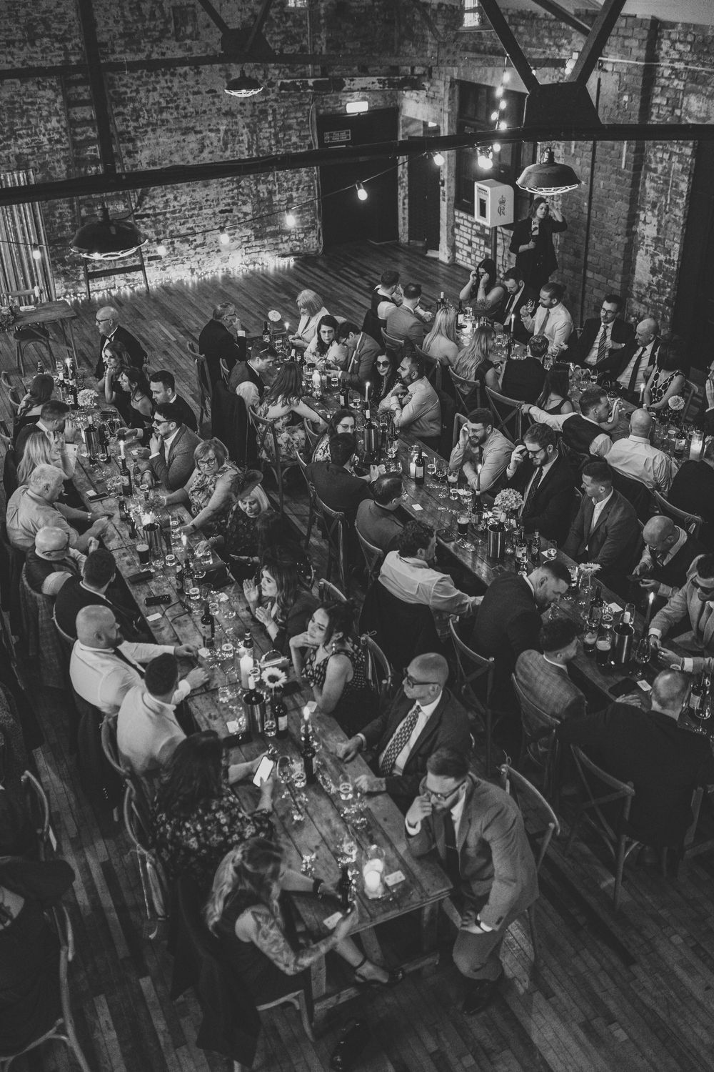 Black and white aerial view of wedding reception guests seated at long tables in rustic brick venue.
