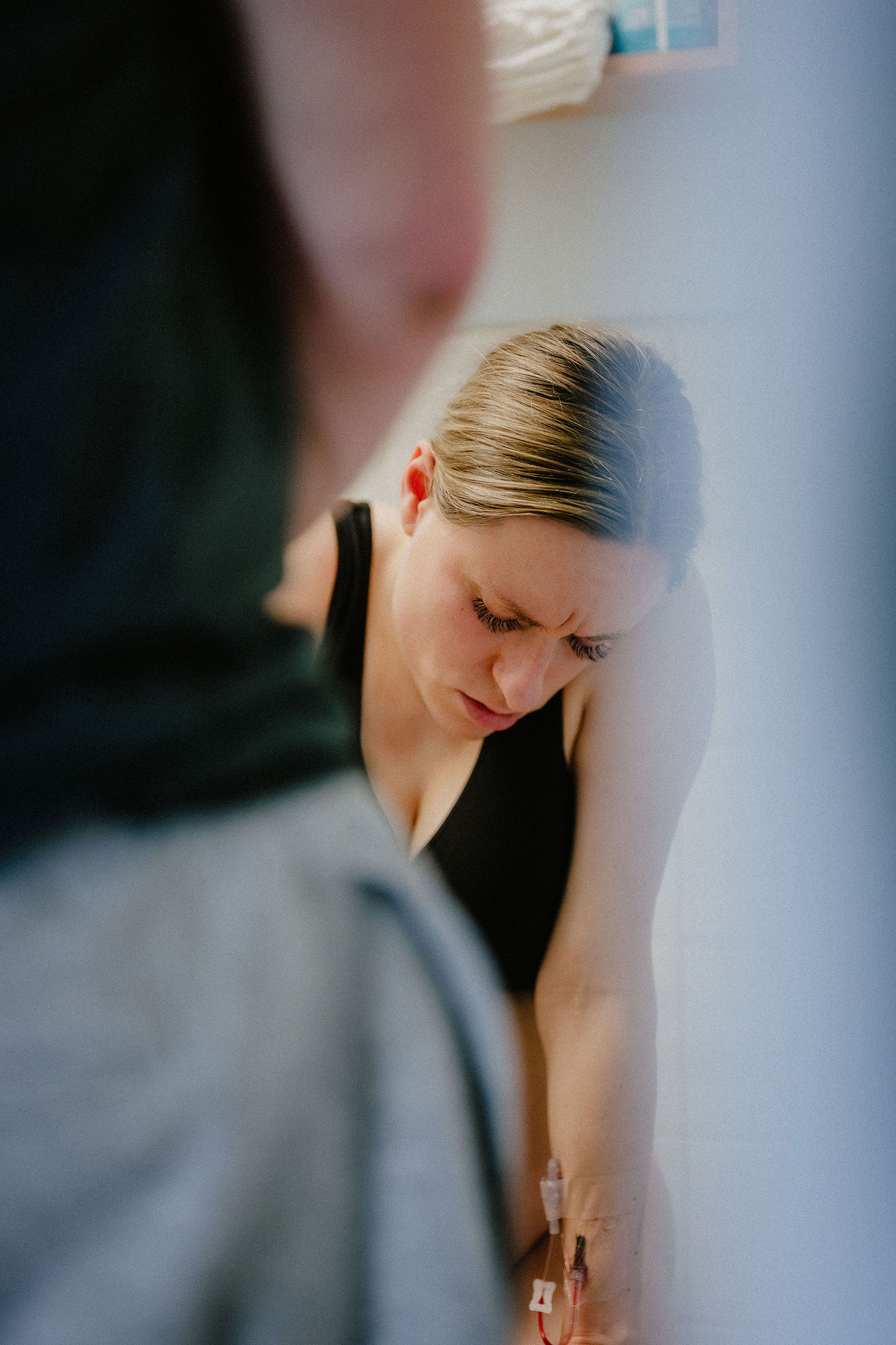 Blonde woman in black tank top leaning against white wall with furrowed brow during labor.