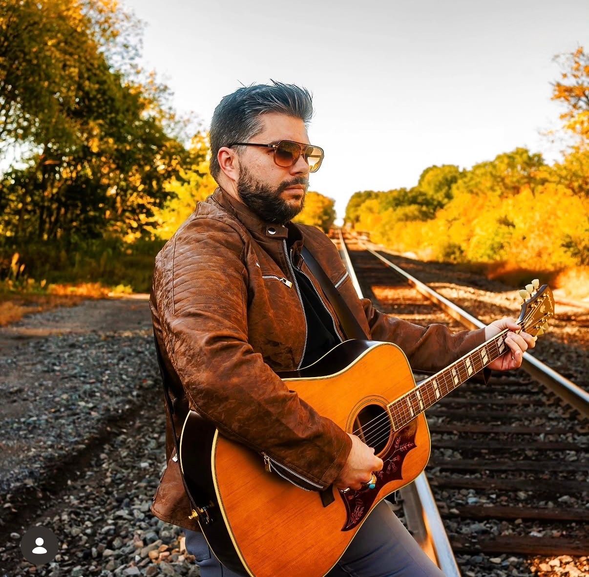 Man in brown leather jacket playing acoustic guitar on railroad tracks surrounded by autumn foliage.