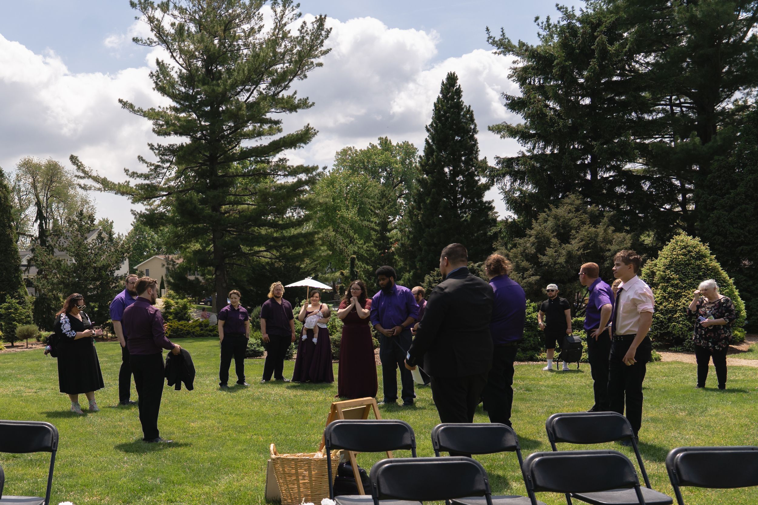 Wedding guests in purple attire gather outdoors on green lawn surrounded by tall trees on sunny day.