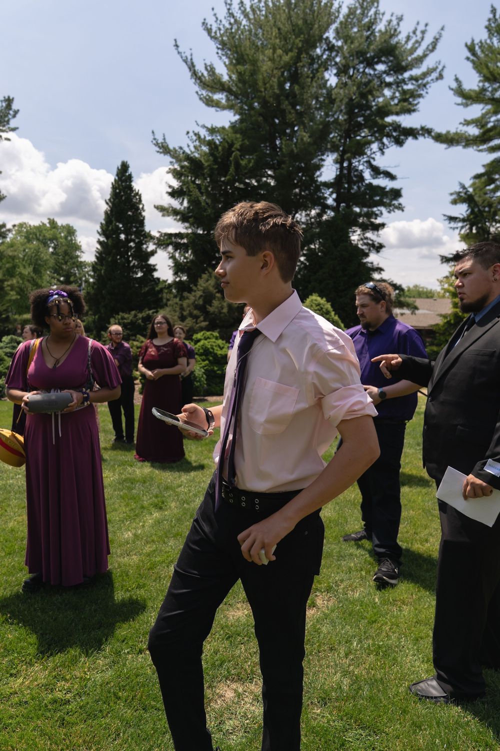 Young man in white shirt and purple tie standing on grass at outdoor wedding celebration.