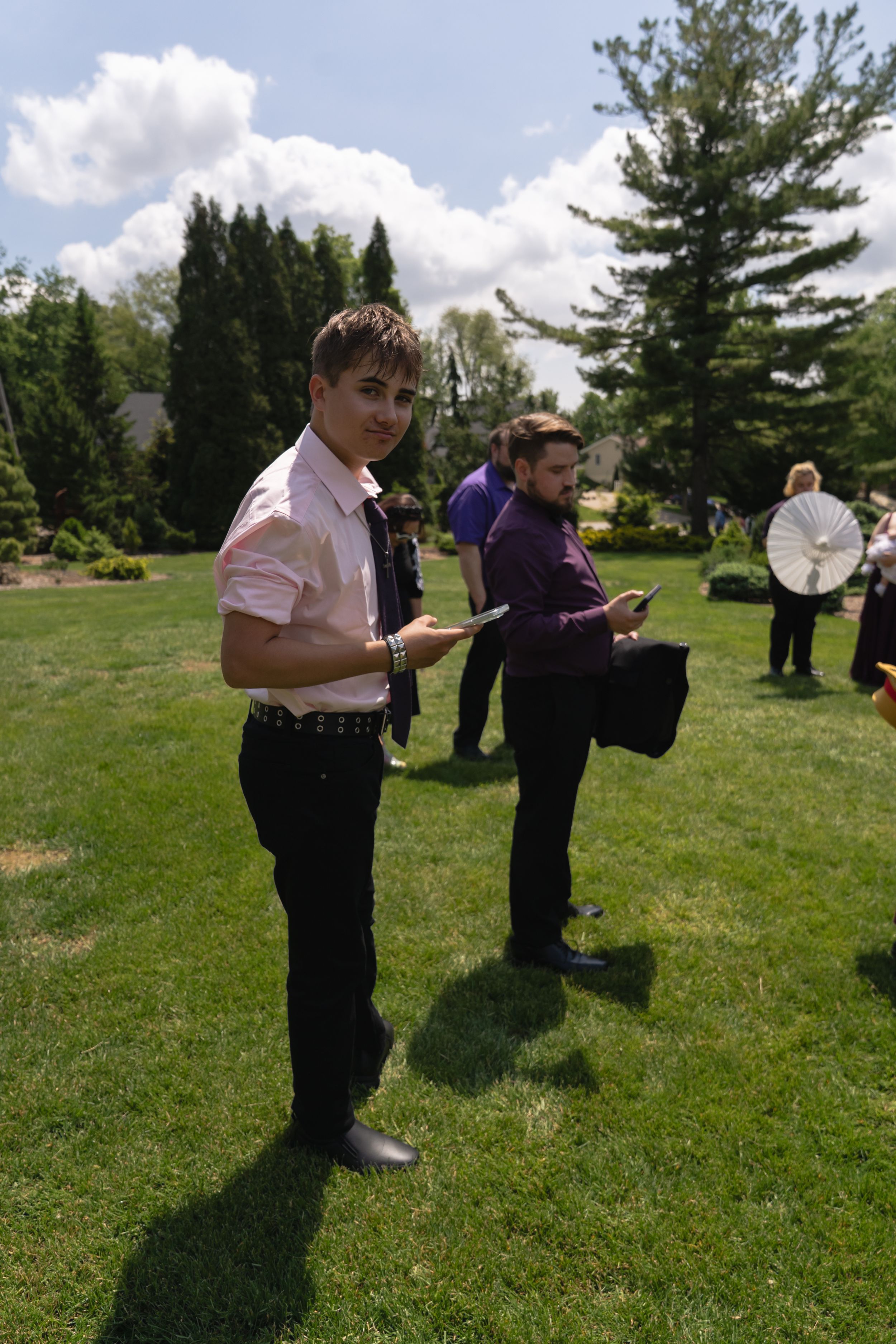 Young man in white shirt and tie holding items stands on green lawn at outdoor wedding event.
