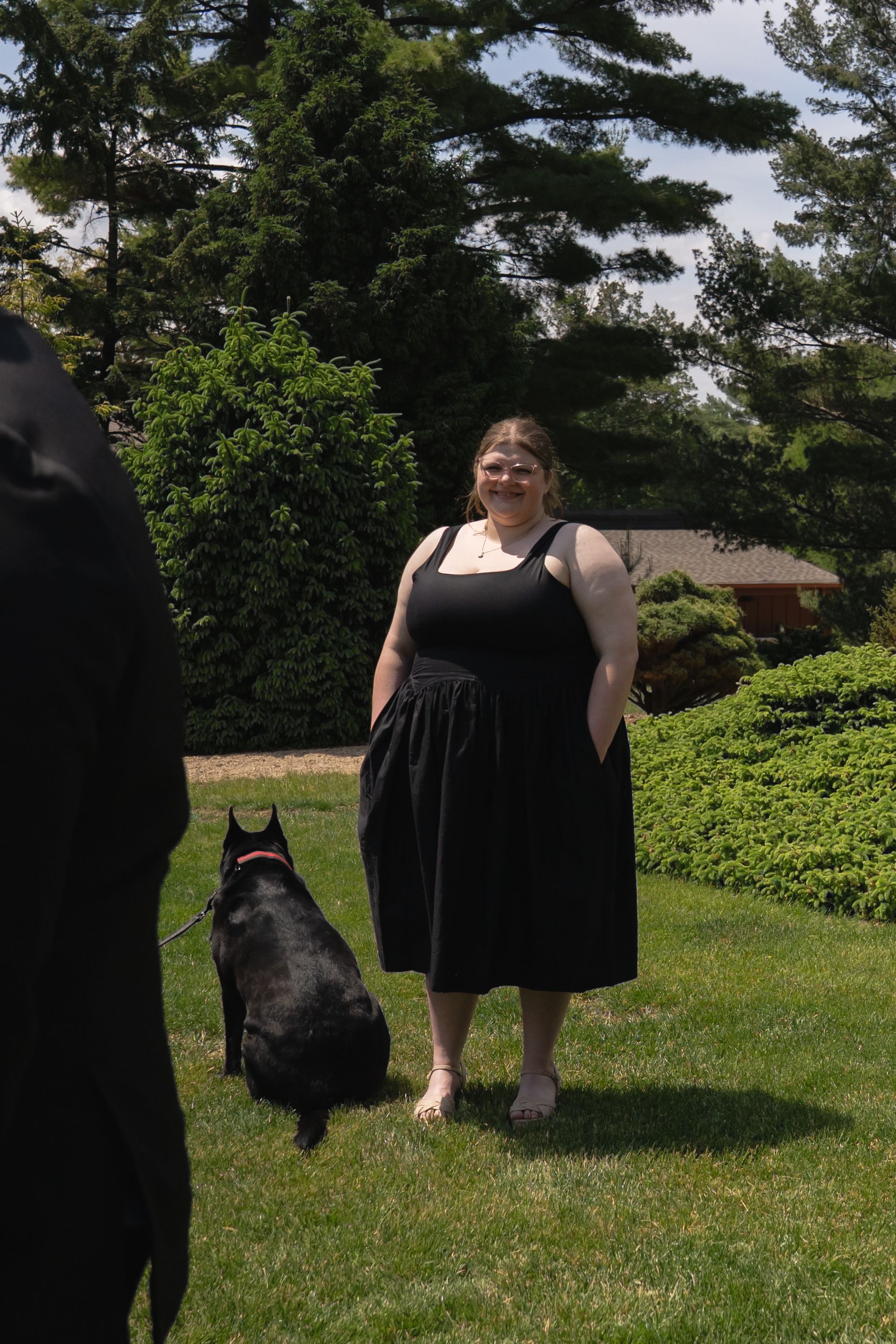 Woman in black dress stands on green lawn with black Labrador dog during outdoor wedding celebration.