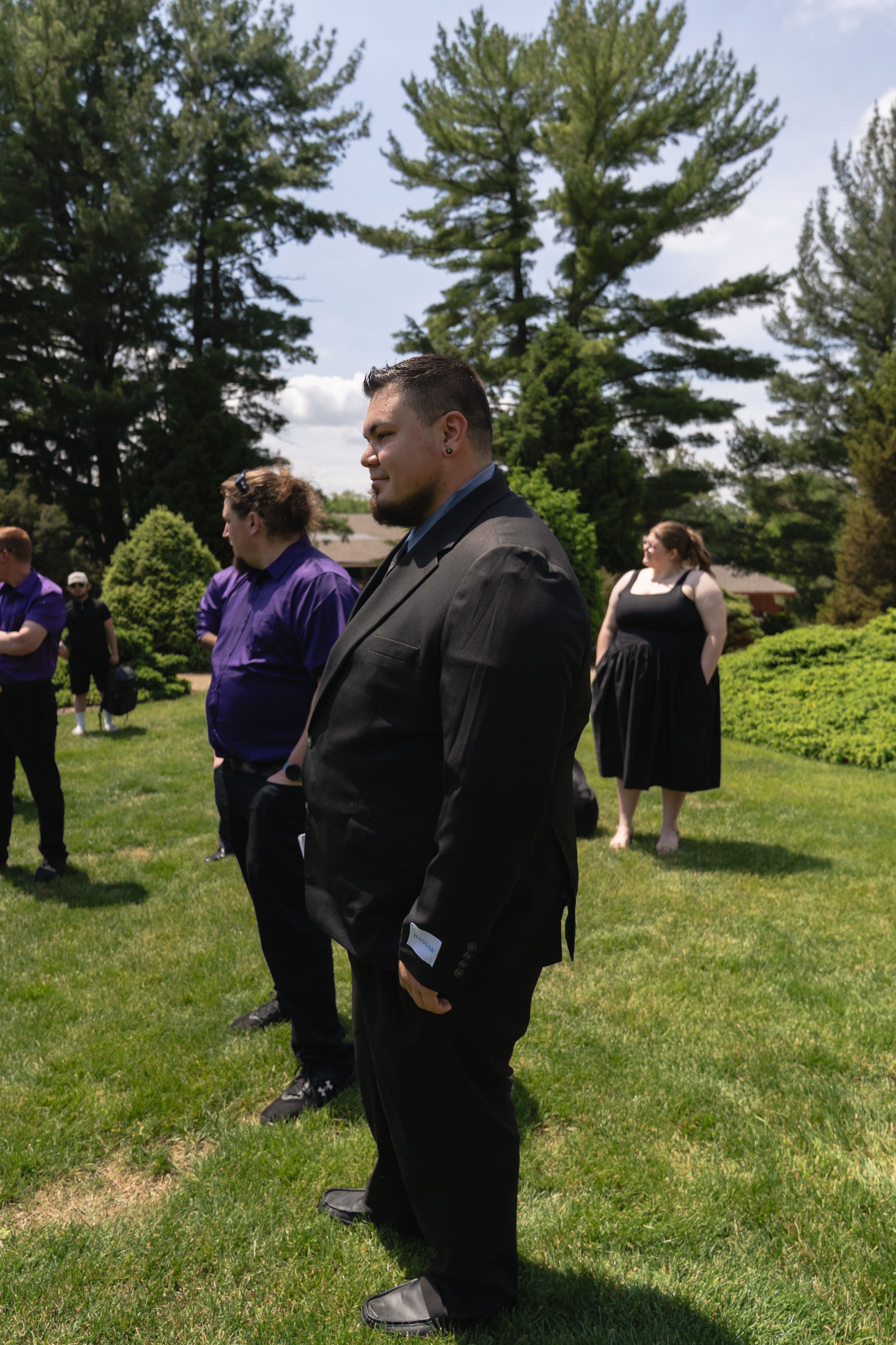 A man in a black suit stands on a green lawn during an outdoor wedding celebration.