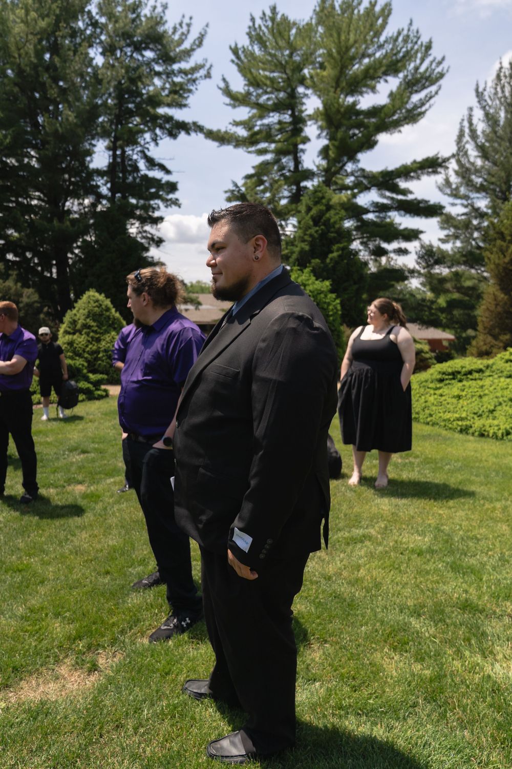 A man in a black suit stands on a green lawn during an outdoor wedding celebration.