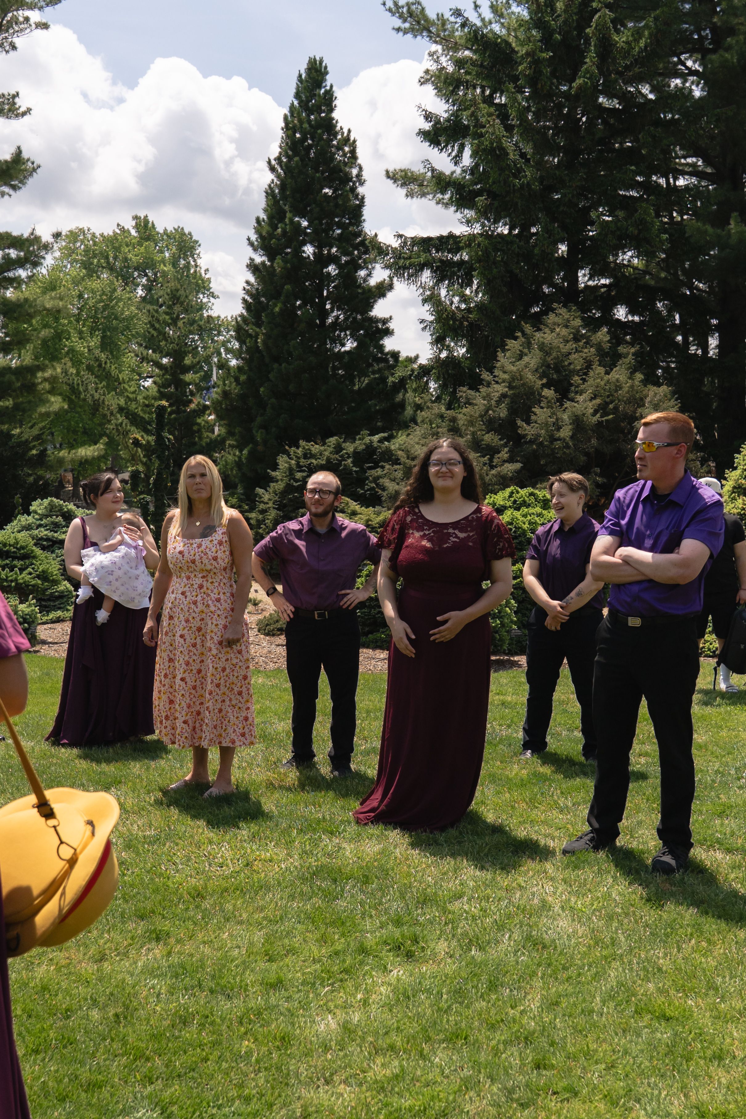 Wedding guests in purple and burgundy attire gather on a sunny green lawn for an outdoor ceremony.