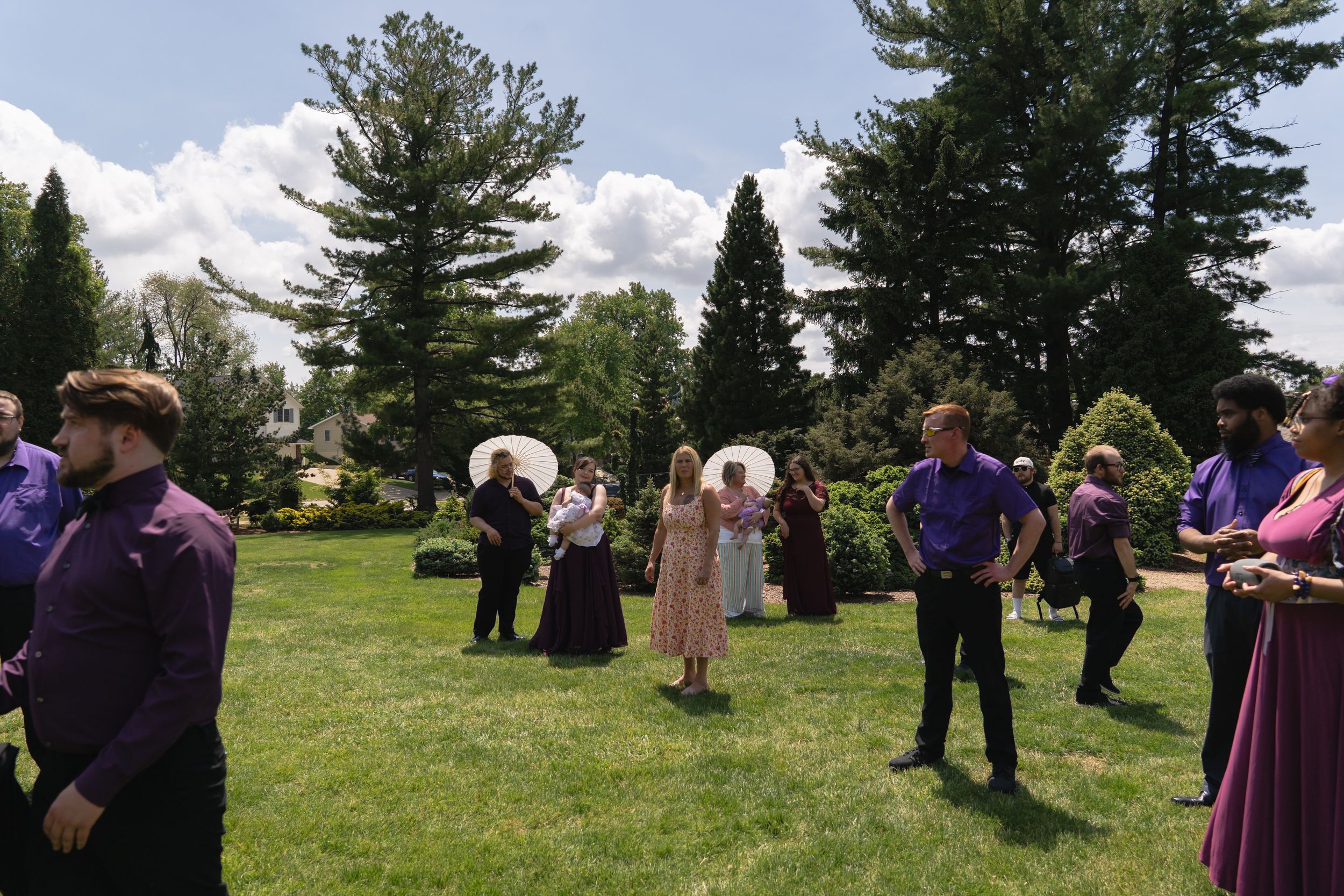 Wedding party in purple attire and guests mingle on sunny green lawn with tall pine trees.